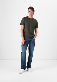 Olivengrønn bomulls t-skjorte med korte ermer, rund hals og en liten logo. Brukt sammen med lys blå slitt jeans og hvite joggesko.