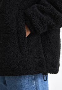 Black fleece jacket with a textured surface, featuring a front pocket, elastic cuffs, and an adjustable hem. Worn over blue jeans.