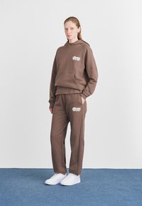 Felpa marrone e pantaloni della tuta coordinati in morbido cotone. Presenta una tasca frontale e un logo stampato in bianco. Indossati con sneakers bianche.
