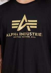 Juoda medvilnė marškinėliai su auksiniu logotipu, kuriame yra stilizuoti sparnai ir raidės "A" bei "I". Tekstas skelbia "ALPHA INDUSTRIES" ir vietovės detales.