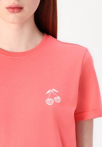 Koral farget bomulls t-skjorte med rund hals og korte ermer, pyntet med hvite broderte kirsebær og ordene "HERRY LOVE."
