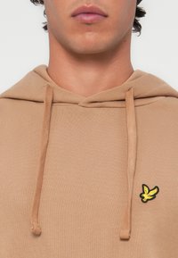Felpa beige con tasca a marsupio, cappuccio regolabile con cordino, polsini a coste e logo ricamato di un uccello nero e giallo sul petto.