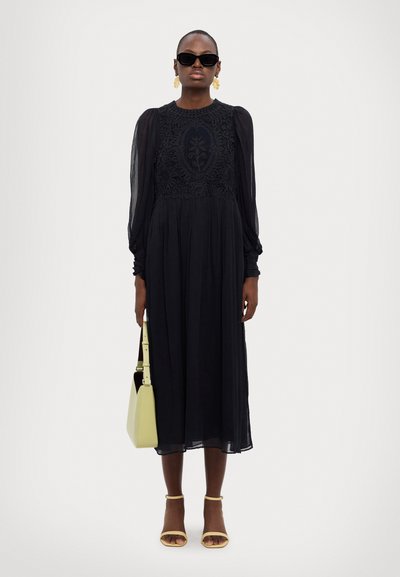 Black long-sleeve dress with floral embroidery at the bodice. Sheer fabric, flowing design. Accessories include a yellow handbag and sandals.