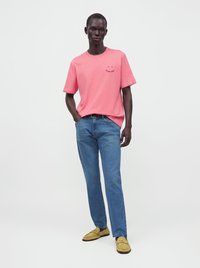 Homme portant un t-shirt rose, un jean bleu et des mocassins jaunes, debout avec une main dans la poche sur un fond uni.