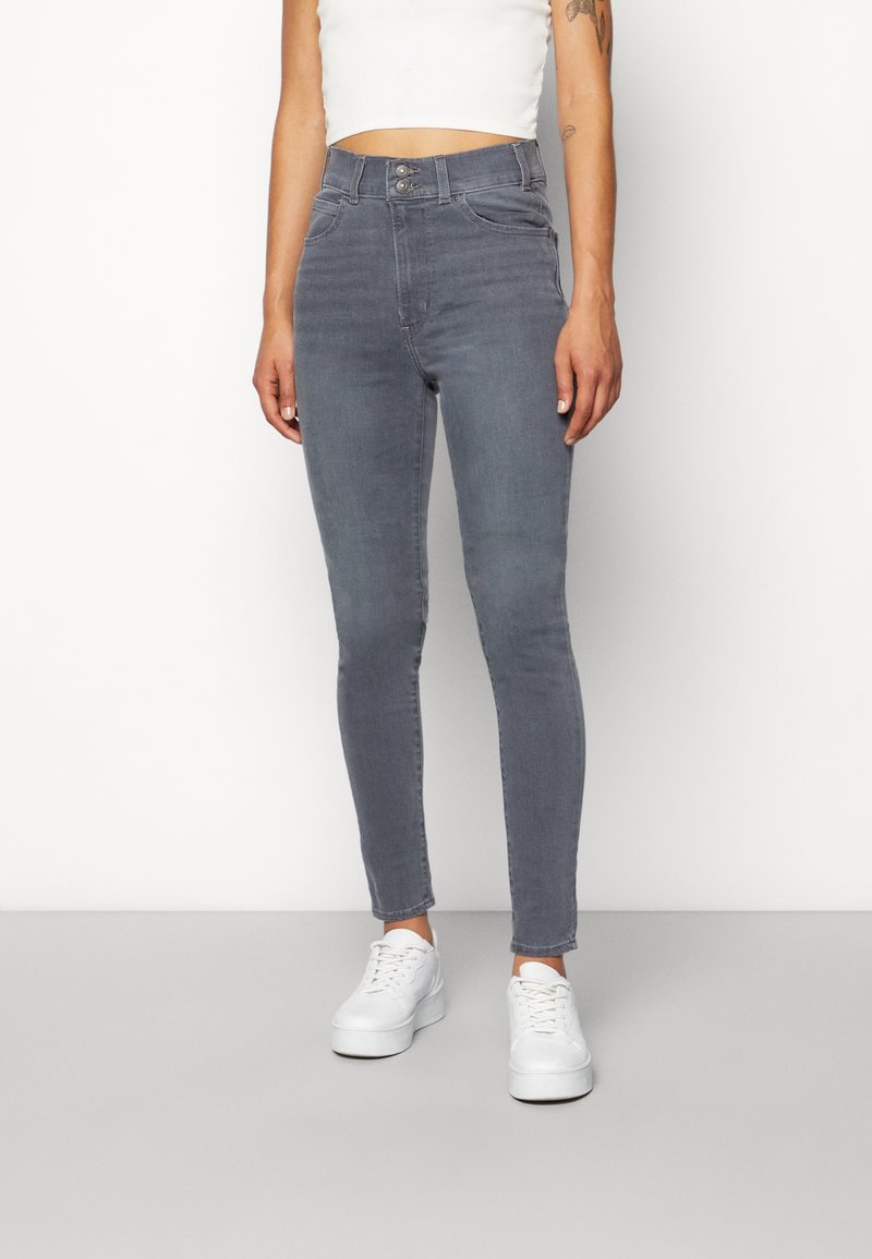 Femme portant un jean gris ajusté taille haute avec double bouton et des baskets blanches, debout devant un fond blanc uni.
