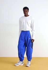 Balta garām piedurknēm blouse, zilas neilona bikses ar melniem akcentiem un baltas kedas ar pelēkiem elementiem. Modelis stāv pret vienkāršu fona.