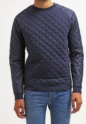 Uomo che indossa una felpa trapuntata blu navy a maniche lunghe abbinata a jeans di denim blu su uno sfondo semplice.