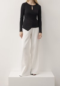 Top nero a maniche lunghe con scollatura a chiave, design arricciato e orlo asimmetrico abbinato a pantaloni bianchi a gamba larga e sandali neri.