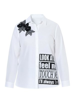 Valkoinen paita, jossa mustaa kukkakirjontaa olkapäällä ja rohkeat mustat tekstit alaosassa. Pitkät hihat, napitettava malli.