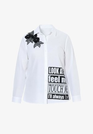 Weißes Hemd mit schwarzer Blumenstickerei auf der Schulter und fettem schwarzem Text auf der unteren Hälfte. Langärmlig, mit Knopfleiste.