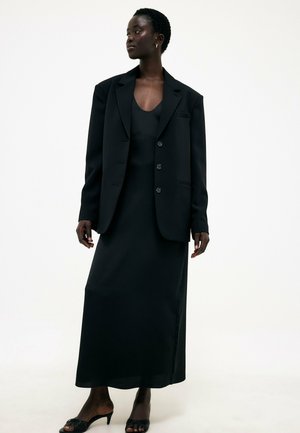 Frau trägt ein langes schwarzes Kleid kombiniert mit einem übergroßen schwarzen Blazer und schwarzen Sandaletten mit Absatz, stehend vor einem schlichten weißen Hintergrund.