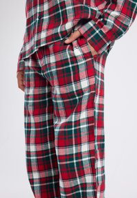 Red, green, and white plaid pajama pants and shirt with a hand casually placed in the pants pocket against a plain background.