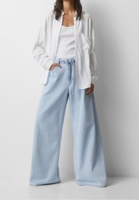 Jeans ampi di colore azzurro chiaro con vita regolabile, abbinati a un top bianco senza maniche e a una camicia bianca lunga con bottoni. Texture liscia.