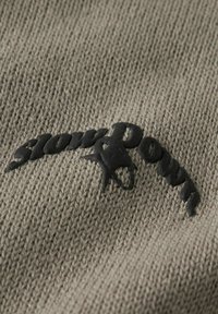 Gros plan sur un tissu gris tricoté avec du texte en caoutchouc noir et un logo en forme d'étoile brodés en arc de cercle.