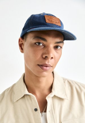 Blue denim cap with a brown leather logo patch, featuring a curved brim and stitching details. Worn with a beige shirt, smooth texture.
