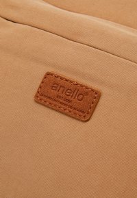 Un tissu beige à la texture lisse présente une étiquette en cuir marron embossée du mot « anello » et des détails de design. L'étiquette est cousue sur le tissu.