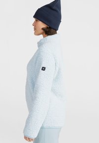 Lichtblauwe, zachte fleece pullover met een hoge kraag, lange mouwen en een klein zwart logo-op-patch op de linkerarm.