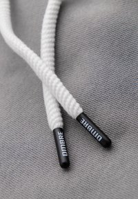 Extrémités de lacets blancs tressés avec embouts en plastique noir portant l'inscription "DMIBRE" sur un fond en tissu gris.