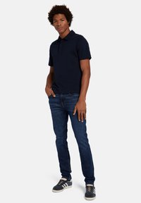 Polo shirt bleu marine en mélange de coton, associé à un jean en denim foncé. Le modèle se tient avec une main dans la poche, portant des baskets foncées à rayures.
