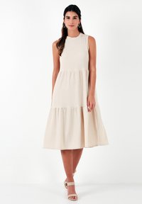 Beige, sleeveless midi dress with a high neckline, gathered tiered skirt, and textured fabric. Worn with light-colored heeled sandals.