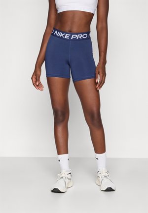 Frau trägt dunkelblaue Nike Pro Shorts, weißes Croptop, weiße Nike Socken und weiße Sportschuhe, steht vor einem schlichten Hintergrund.