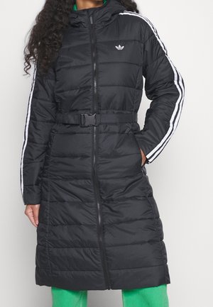 Femme portant un long manteau matelassé noir Adidas avec des rayures blanches sur les manches et une ceinture à la taille, associé à un pantalon vert.