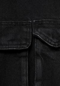 Zwarte denim stof met een gladde textuur, donkere stiksels en opgestikte zakken op de borst. De indeling is eenvoudig en functioneel.