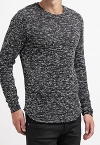 Muž v dlouhém rukávu, černobílé melírované pletené svetru s kulatým výstřihem a černých kalhotách, stojící s uvolněnýma rukama podél těla.