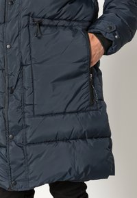 Marinblå puffjacka med quiltat mönster, försedd med tryckknappar, en ficka med dragkedja och elastiska ärmslut för värme och komfort.