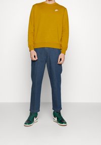 Gul crewneck sweatshirt med vitt logotyp, tillsammans med blå raka jeans och gröna hög-top sneakers med vita detaljer.