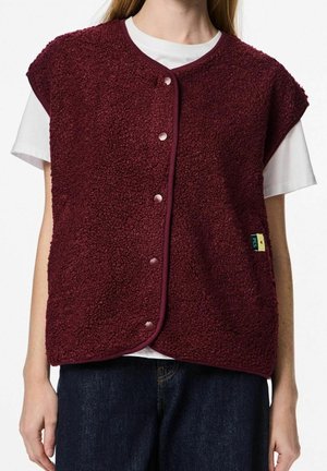 Persona vistiendo un chaleco de forro polar sin mangas color burdeos con botones sobre una camiseta blanca de manga corta y jeans azul oscuro, de pie frente a un fondo blanco.