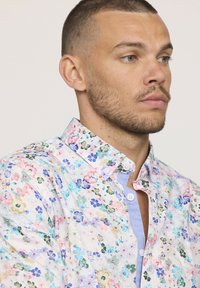Chemise à motifs floraux avec une base blanche, ornée de petites fleurs multicolores. Design à boutons avec un col intérieur bleu clair.