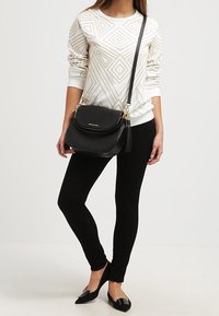 Sort crossbody taske med guldfarvede detaljer, frynseaccent og afrundet design. Båret sammen med en hvid sweater med geometrisk mønster og sorte leggings.