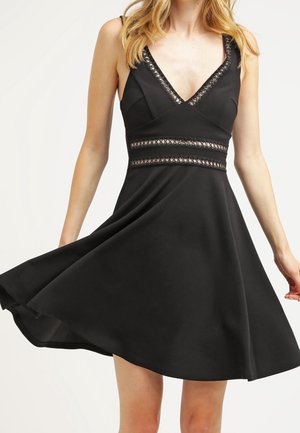 Kobieta w czarnej sukience bez rękawów z koronkowym obszyciem na dekolcie w kształcie litery V i w talii, obracająca się, aby rozłożyć spódnicę.