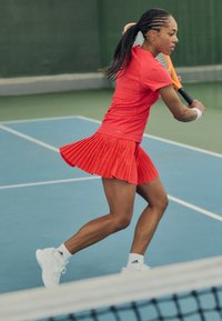 Red pleated tennis skirt and matching fitted top, with white athletic shoes, on a blue court during play.