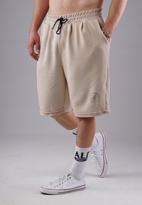 Beige cotton blend shorts with an elastic waistband, black drawstring, side pockets, and a logo detail near the hem. Worn with white shoes and socks.