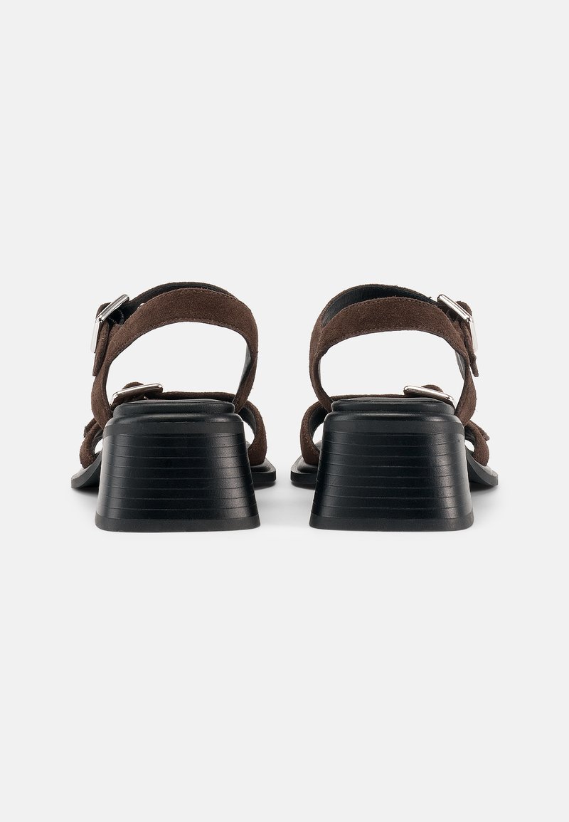 Paar braune Wildleder-Sandalen mit offenen Zehen, schwarzen Blockabsätzen und verstellbaren silbernen Schnallen, von hinten auf weißem Hintergrund betrachtet.