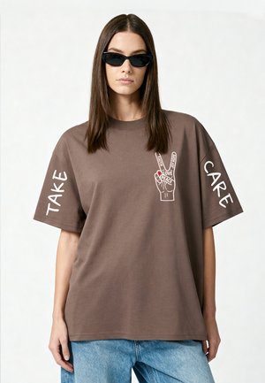 Femme portant des lunettes de soleil et un t-shirt marron oversize avec "Take Care" sur les manches et un motif du signe de la paix avec les mots amour, foi, joie, espoir.