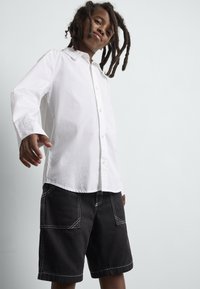 Camicia bianca a maniche lunghe con colletto, abbinata a pantaloni corti neri con cuciture bianche e due tasche frontali.