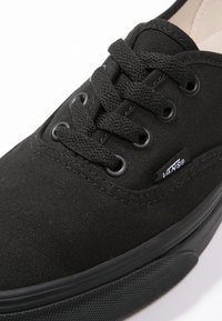 Baskets en toile noire avec embout rond en caoutchouc, lacets plats passant par six œillets, côté texturé et une étiquette avec le logo Vans.