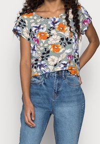 Svetle zelené tričko s krátkými rukávy a květinovým vzorem, na kterém se objevují oranžové, fialové a modré květiny, v kombinaci s džínovými kalhotami s vysokým pasem v modré barvě.