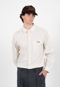 Homme portant une chemise beige à fines rayures avec le mot "Interdit" sur la poche et une casquette marron avec le texte "L'art d'être en retard", mains dans les poches.