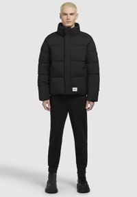 Schwarze Daunenjacke mit hohem Kragen, ausgestattet mit horizontalem Steppmuster, Frontreißverschluss und Logopatch. Getragen mit schwarzen Hosen und Stiefeln.