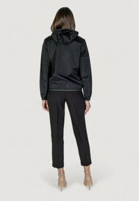 Giacca nera, leggera, con cappuccio, caratterizzata da una texture liscia e polsini elasticizzati. Indossata sopra pantaloni neri sartoriali e scarpe color carne.