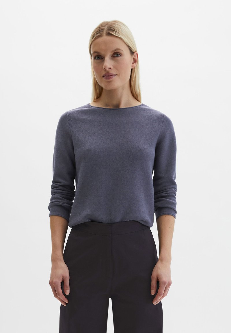 Woman with straight blonde hair wearing a long-sleeve gray sweater and high-waisted black pants, standing against a white background.