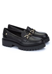 Mocasines de piel negra con una suela gruesa negra, que cuentan con un acento de metal dorado y una textura suave. Diseño clásico con punta redondeada.