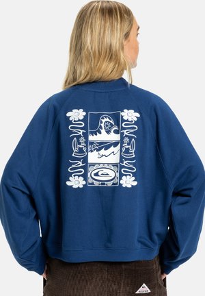 Blond persoon met de rug naar voren, draagt een blauw sweatshirt met een witte afbeelding van golven, een surfplank, bloemen en keramische potten op de achterkant.