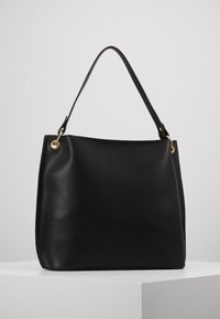 Czarna skórzana torebka z pojedynczym paskiem przymocowanym złotymi pierścieniami, prezentowana na białym postumencie na tle jednolitego szarego tła.