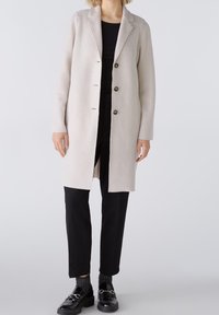 Abrigo de lana gris claro con solapa de muesca, cuatro botones frontales y mangas largas, combinado con pantalones negros y zapatos brillantes.