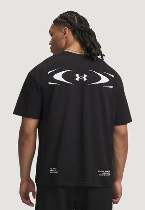 Uomo con i capelli intrecciati che indossa una t-shirt nera con un grande logo bianco Under Armour e testo sulla parte superiore della schiena e sull'orlo inferiore.